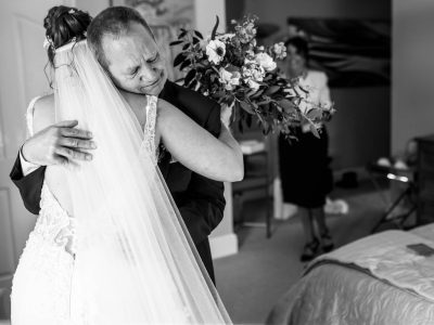 bride getting ready 20 In an emotional scene, as the bride getting ready embraces a man tightly in a lace gown, a woman holds flowers in the background. The moment is intimate and beautifully captured by a second shooter.