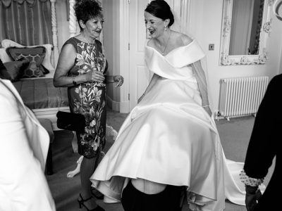 bride getting ready 22 The second shooter captures a bride in a satin gown laughing as she gets ready, with others around her. A person hides under her dress while another woman in a floral dress chats nearby, capturing this joyful moment beautifully.
