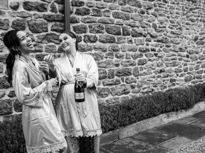 bride getting ready 28 Two photographers capture the joyous moment as two women in robes laugh, holding a large bottle of champagne against a stone wall.