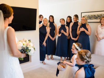 bride getting ready 33 A bride in a veil holds flowers, facing eight bridesmaids in navy dresses and two flower girls, while the second shooter captures the serene moment. One woman in white stands beside them in a room with a TV and framed picture.