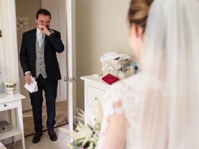bride getting ready 35 In a heartfelt moment, the bride stands with her back to the camera, as the groom, captured by two photographers during preps, wipes a tear in an open doorway.
