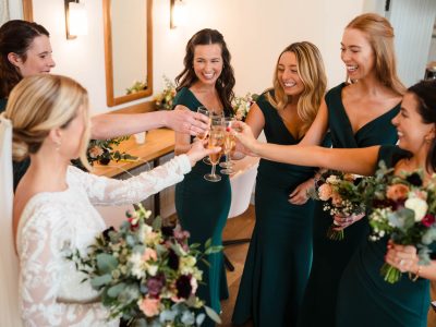 bride getting ready 36 A bride and five bridesmaids in dark green dresses toast with champagne glasses, holding bouquets, and smiling in a well-lit room as the second shooter captures every joyful moment.