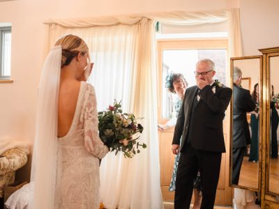 bride getting ready 37 A bride in a white dress stands facing her emotional father, while a woman smiles behind him. In the beautifully draped room with a sofa and large mirror, two photographers capture groom and bride preps seamlessly.