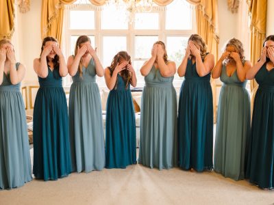 bride getting ready 39 Eight women in teal and blue dresses stand side by side, covering their eyes with their hands, in a room with large windows and ornate yellow curtains, as the second shooter captures the bride getting ready.