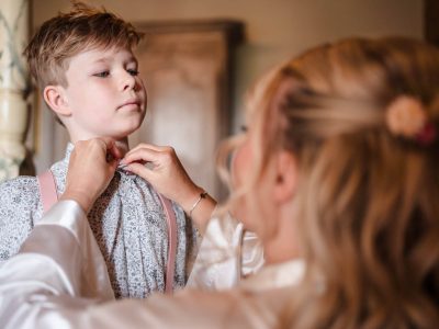 bride getting ready 43 A young person adjusts a bowtie on another child, who is wearing a floral shirt and suspenders, reminiscent of a candid moment as if captured by a second shooter. The setting appears indoors with wooden furniture in the background.