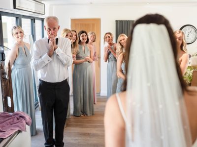 bride getting ready 44 A man in a white shirt looks surprised as he sees a woman in a bridal gown. Bridesmaids in gray dresses stand smiling, while the second shooter captures the joyous scene of the groom and bride's preps unfolding.
