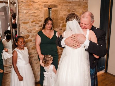 bride getting ready 47 A man in a kilt hugs a bride wearing a veil as the second shooter captures the moment. Nearby, two women, a girl, and a child with a floral headpiece stand against the textured stone wall with a mirror, adding to the charm of this wedding scene.