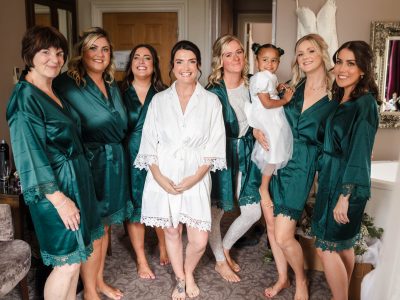 bride getting ready 48 As two photographers capture groom and bride preps, a bride in a white robe is surrounded by bridesmaids in green robes, standing in a room. Meanwhile, the second shooter captures one bridesmaid holding a young girl in a white dress.