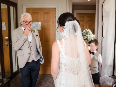 bride getting ready 49 The bride, caught in a moment straight out of a fairytale, stands facing an older man who looks surprised, covering his mouth. Meanwhile, two photographers capture groom and bride preps. Further in the room, two children in suits are visible, one holding flowers.