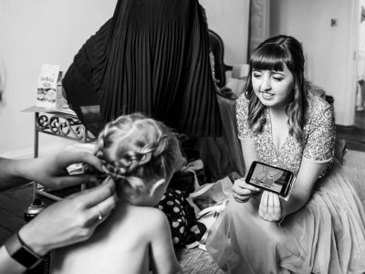 bride getting ready 6 A woman shows a smartphone to a child with styled hair, capturing the moment as another person adjusts the child's braid. The behind-the-scenes action unfolds like a scene of bride getting ready, each detail attended to with care and precision.