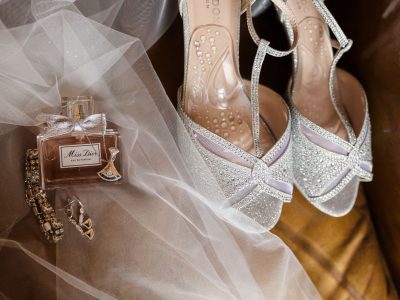 bride getting ready details Perfume bottle, glittery high-heeled shoes, and jewelry on a brown surface with a sheer fabric overlay.