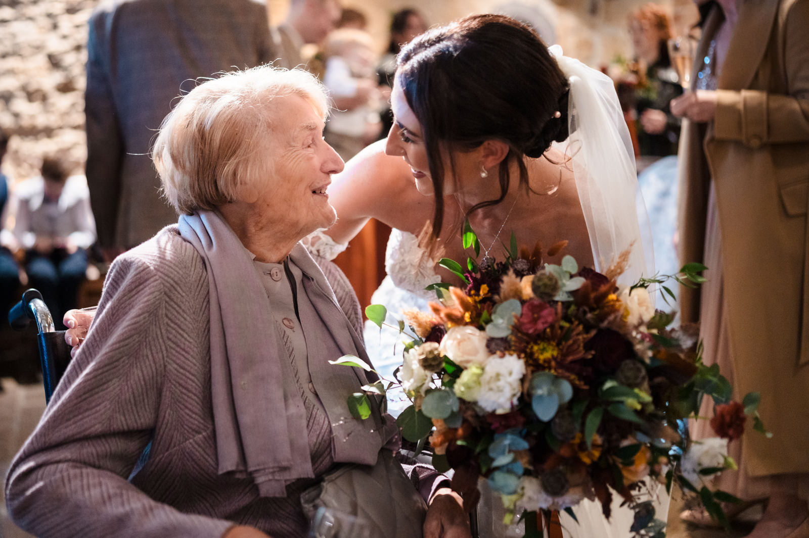 A bride in a white dress and veil beams as she kneels beside an elderly woman in a wheelchair, clutching a colorful bouquet. Captured with the essence of documentary wedding photography, the candid scene unfolds indoors amid softly blurred onlookers.