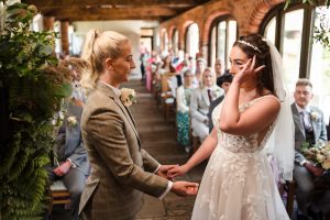 Two people exchange vows in a heartfelt ceremony at Dodmoor House. One, in a sharp suit, gently holds the other's hand, who is gracefully adorned in a white dress with a veil. Guests sit attentively in the background, ready to cherish these Dodmoor House photos forever.