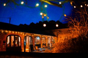 A cozy, warmly lit stone building with arched windows and string lights in a nighttime setting, surrounded by trees and shrubs. Capture the enchanting charm of Dodmoor House Northamptonshire venue as it effortlessly transforms any event into a magical experience.