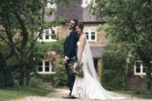 The bride and groom stand back-to-back on a tree-lined path in front of Dodmoor House, a serene Northamptonshire venue. The bride holds a bouquet and wears a white dress while the groom sports a dark suit, creating picture-perfect Dodmoor House photos.
