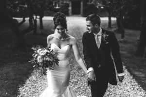 A couple in wedding attire walks hand in hand down a gravel path, surrounded by trees at Dodmoor House. The bride holds a bouquet of flowers. Captured by a skilled Dodmoor House wedding photographer, the image is elegantly presented in black and white.