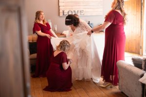Bride in a white gown surrounded by three women in red dresses adjusting her train, with a "LOVE" sign on the wall behind them at the charming Dodmoor House Northamptonshire venue.
