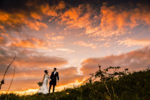 A couple in wedding attire stands on a grassy hill at sunset, holding hands beneath a sky filled with dramatic orange and pink clouds—capturing the essence of Dodmoor House Wedding Photography.