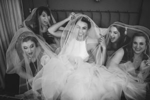 Five women are laughing and playfully covering themselves with a wedding veil while sitting on a bed at Dodmoor House, the best wedding venue in Northamptonshire.