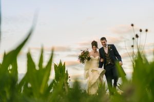 A bride and groom walk happily through a field of tall grass, the sky and clouds beautifully complementing their joy. Capturing these moments, a Dodmoor House Wedding Photographer ensures every emotion is preserved in stunning detail.
