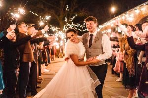 Bride and groom smile as guests hold sparklers, crafting a lit pathway at Dodmoor House, the enchanting Northamptonshire venue. The bride in a fur stole and gown, alongside the groom in a vest, stand amidst twinkling lights captured beautifully by the Dodmoor House Wedding Photographer.