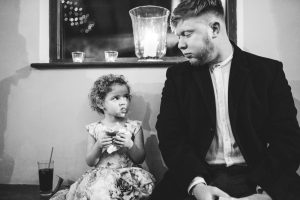 A young girl with a cupcake and a man sit together on a bench. With frosting smudged on her face, they share a warm gaze. In the background, where Dodmoor House is often captured in wedding photography, a lit candle flickers softly.