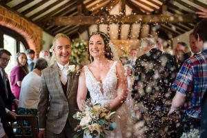 A newlywed couple stands smiling indoors at the beautiful Dodmoor House Northamptonshire venue, surrounded by guests. Confetti is being thrown. The bride holds a bouquet and wears a white dress; the groom in a checked suit beams with happiness, perfectly captured in stunning Dodmoor House wedding photography.