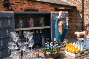 A person adjusts a sign inside an outdoor bar setup, complete with a variety of bottles and two-tiered stands with empty glasses, capturing the essence of Dodmoor House Wedding Photography at this timeless venue.