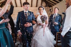 The bride and groom, in elegant formal attire, walk down the aisle at Dodmoor House in Northamptonshire, beaming as guests shower them with confetti in the beautifully decorated indoor setting.