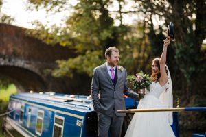 Bride and groom stand joyfully on a blue canal boat, with the bride raising a hat and holding a bouquet. A stone bridge and trees create a picturesque backdrop, reminiscent of the charming scenes found in Dodmoor House photos from Northamptonshire's best wedding venue.
