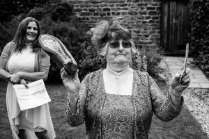 An older woman in a lace dress and hat holds a shoe and paper, captured beautifully by Dodmoor House Wedding Photography. In the background, a smiling woman also holds paper as they stand outside beside the charming stone building.