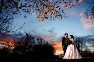A bride and groom stand together outdoors at sunset, surrounded by bare trees and orange leaves, under a colorful sky—a perfect moment captured in Dodmoor House Wedding Photography.
