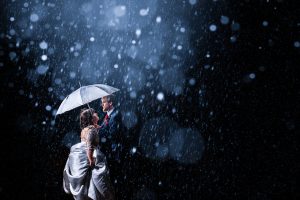 A couple stands under an umbrella in the falling rain at Dodmoor House, their love illuminated by soft light against the dark background, capturing a moment that could grace any Dodmoor House Wedding Photography album.