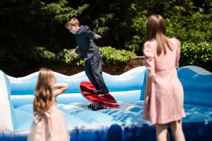 At Dodmoor House, a boy in a suit rides a mechanical surfboard on an inflatable, with two girls in dresses watching him. The scene is as delightful as any moment captured by a Dodmoor House Wedding Photographer.