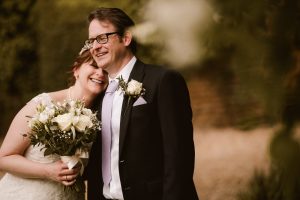 A bride and groom are captured in a tender moment at Dodmoor House, Northamptonshire. She rests her head on his shoulder, both smiling warmly while holding a bouquet of white flowers at this stunning venue, perfect for any Dodmoor House wedding photographer's portfolio.
