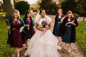 A bride in a white dress and veil strolls outside Dodmoor House with four bridesmaids holding bouquets, smiling and chatting. They're on a gravel path framed by lush greenery, capturing perfect moments for Dodmoor House photos at Northamptonshire’s best wedding venue.