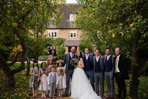 The wedding party gathers outdoors at the picturesque Dodmoor House, with the bride, groom, bridesmaids, groomsmen, and children in formal attire. A charming brick house and lush trees frame this memorable moment at Northamptonshire's best wedding venue.