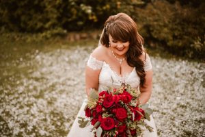 At a Dodmoor House wedding, the bride in her white gown holds a bouquet of red roses. She stands on grass scattered with petals, smiling and looking down. Perfect for Dodmoor House wedding photography, this moment reflects pure joy and romance.