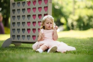 A girl in a pink dress sits on the grass blowing bubbles in front of a game board with heart-shaped cutouts. This whimsical scene captures the charm often found at Dodmoor House weddings, a picturesque Northamptonshire venue perfect for capturing enchanting moments.