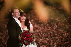 A couple in wedding attire smiles while standing on fallen leaves at Dodmoor House, renowned as the best wedding venue. The bride holds a bouquet of red roses and greenery, capturing a perfect moment for Dodmoor House Wedding Photography.