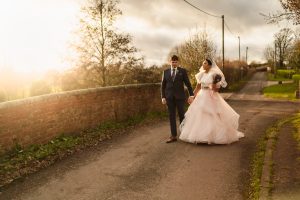 A couple in wedding attire walks hand in hand along a rural road near a brick wall, with trees and a grassy landscape under a cloudy sky, reminiscent of the picturesque charm captured by Dodmoor House Wedding Photography.