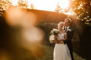 A couple in wedding attire stands embracing and smiling under a sunlit archway at Dodmoor House, Northamptonshire's best wedding venue, with trees and grass providing a picturesque background.
