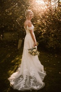 The bride, in a white lace gown, stands gracefully on the grass at Dodmoor House. Sunlight filters through the trees as she holds her bouquet, creating a perfect moment for Dodmoor House wedding photography.