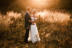 In a sunlit field near Dodmoor House, the bride and groom embrace amidst tall grass. The groom's suit contrasts beautifully with the bride's white dress, as they are bathed in warm, golden light. This idyllic moment captured by a talented Dodmoor House wedding photographer highlights their special day.
