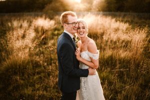 Amidst a sunlit field with tall grass and a warm, golden glow, a couple in formal attire embraces and smiles. This enchanting scene could easily belong to the magical Dodmoor House, renowned for its breathtaking wedding photography opportunities.
