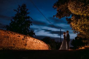 A bride and groom stand facing each other at night near a brick wall, silhouetted against a dark blue sky with scattered clouds and stars, surrounded by trees—a picturesque moment captured perfectly through Dodmoor House Wedding Photography.