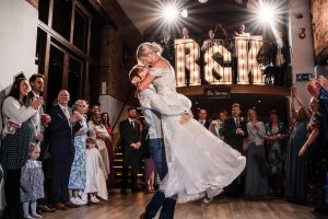 Bride and groom share a kiss during their first dance at Dodmoor House, a warmly lit venue surrounded by guests. Large illuminated initials are displayed in the background, perfectly captured as part of Dodmoor House Wedding Photography.