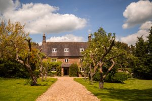 A stone house with a gabled roof and multiple chimneys, Dodmoor House in Northamptonshire is surrounded by trees and bushes, with a gravel path leading to the entrance under a partly cloudy sky. Known as the best wedding venue, it offers a picturesque setting for unforgettable photos.