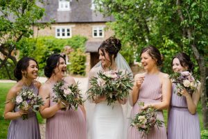 Five women in pastel dresses, holding flower bouquets, stand smiling in a garden framed by greenery, perfectly capturing the essence of a Dodmoor House wedding. The charming brick building behind them highlights why it's considered the best wedding venue.