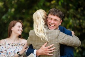 A smiling man in a suit hugs a blonde woman outdoors, while a woman in a floral dress looks on, all captured beautifully by Dodmoor House Wedding Photography.